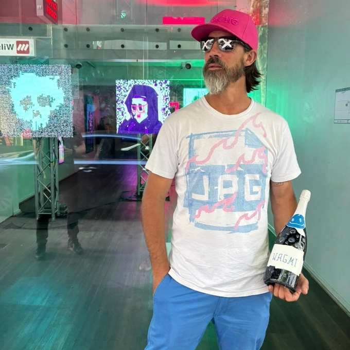 A bearded man wearing a pink 'TRUMP' cap, sunglasses, and a white t-shirt with a 'JAG' design holds a small black device labeled 'WAGMI' in what appears to be an art gallery with neon-lit digital displays.