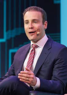 A man in a dark suit with a pink striped tie speaks while seated on stage, wearing a headset microphone.