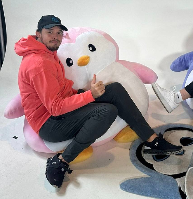 A man wearing a black cap, pink hoodie, and black pants sits on a large pink and white plush penguin, giving a thumbs-up. He wears black On sneakers, and there's a small blue plush and a character rug visible nearby.