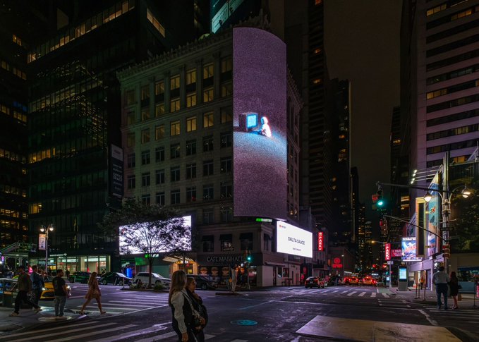 A nighttime cityscape of a busy street intersection, likely in New York City, featuring tall buildings with illuminated billboards, including a large vertical display and one for 'DELTA LABS.'
