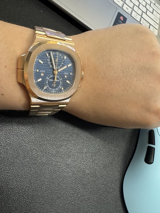 A wrist wearing a rose gold Patek Philippe Nautilus watch with a blue dial and a perpetual calendar complication, resting near a laptop keyboard and a blue ergonomic mouse.