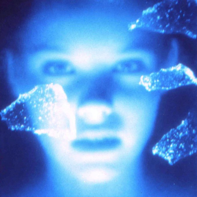 An artistic, blue-tinted portrait of a face partially obscured by glowing, crystalline shapes held in front of it, creating a mystical or futuristic aesthetic.