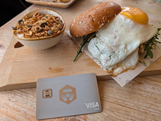 A breakfast scene with a sesame bun egg sandwich, a bowl of granola with pumpkin seeds, and a Visa Debit metal card on a wooden board.