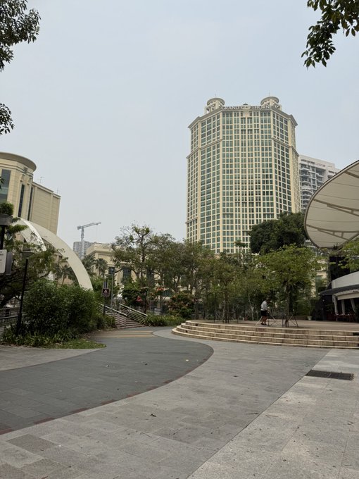 Urban plaza view with a tall yellow and white residential high-rise building in the background, surrounded by trees and paved walkways.