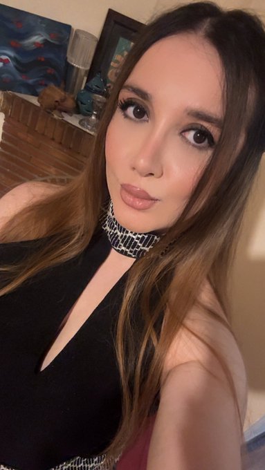 A woman with long brown hair takes a selfie wearing a black top and a patterned choker, with a painting and lamp visible in the background.