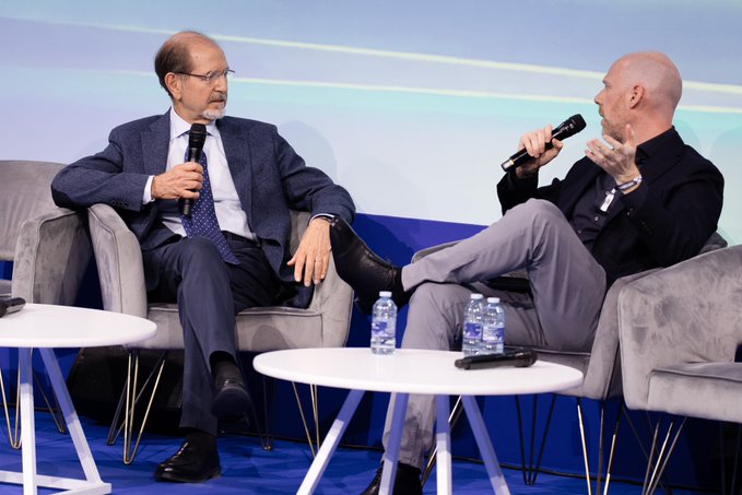 The image shows two men sitting at a table with microphones, engaged in conversation. The man on the left is wearing glasses and a suit, while the other man has a bald head and is dressed more casually. They are seated on chairs placed around a round white table. In front of them, there are several water bottles and a cell phone.

The image also contains text that reads "Can a blockchain be fully secure without a native token at its core? According to Turing Award winner @silviomicali, the answer is a resounding yes #Blockchain #Crypto #FiatChain". This suggests that they might be discussing the security and potential applications of blockchain technology.