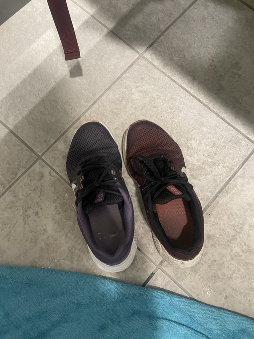 A pair of mismatched Nike shoes—one dark blue/purple and one dark red/maroon—sitting on a tiled floor, with a teal towel visible at the bottom of the frame.
