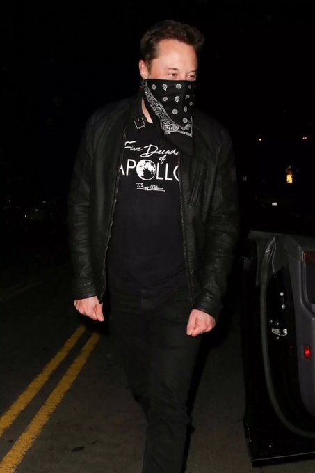 Elon Musk walks at night wearing a black bandana over his face, a black leather jacket, and a black t-shirt with 'Five Despacito APOLLO' text, near a Tesla vehicle.