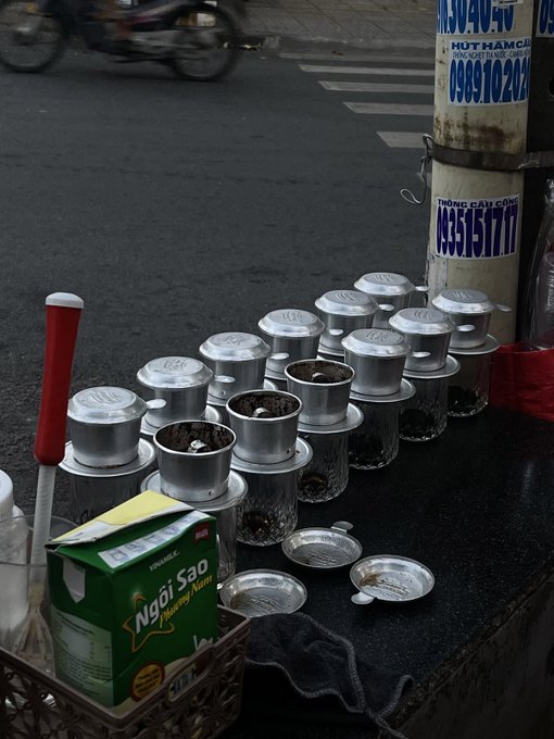 A Vietnamese street coffee setup with metal phin filters stacked on glasses containing ground coffee, alongside a green box of 'Ngôi Sao' powdered creamer, with a motorbike passing on the street and phone numbers visible on a sign in the background.