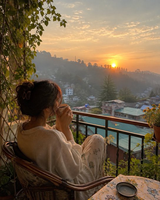 A young woman with a butterfly hair clip sits on a balcony, drinking from a cup while watching a golden sunrise over a hillside town.