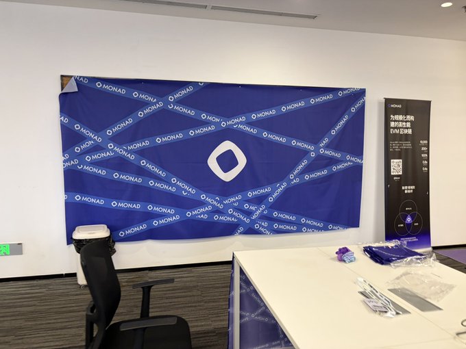 The image features a large blue banner with white text, which appears to be a promotional or advertising sign for Monad. The banner is displayed prominently against a wall, possibly in an office setting. In front of the banner, there are two chairs - one black and one gray. A desk can also be seen nearby, suggesting that this might be a workspace or meeting area related to Monad projects.