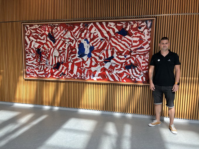 A man in a black polo shirt and shorts stands next to a large framed artwork depicting abstract red, white, and blue patterns, mounted on a wooden slatted wall.