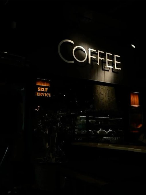 An illuminated 'COFFEE' sign glows in the darkness above a self-service coffee station, with a smaller 'SELF SERVICE' sign visible below.