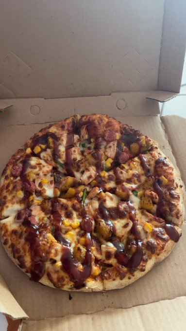 A close-up photograph of a pizza topped with barbecue sauce, cheese, corn, and meat chunks inside a cardboard delivery box.