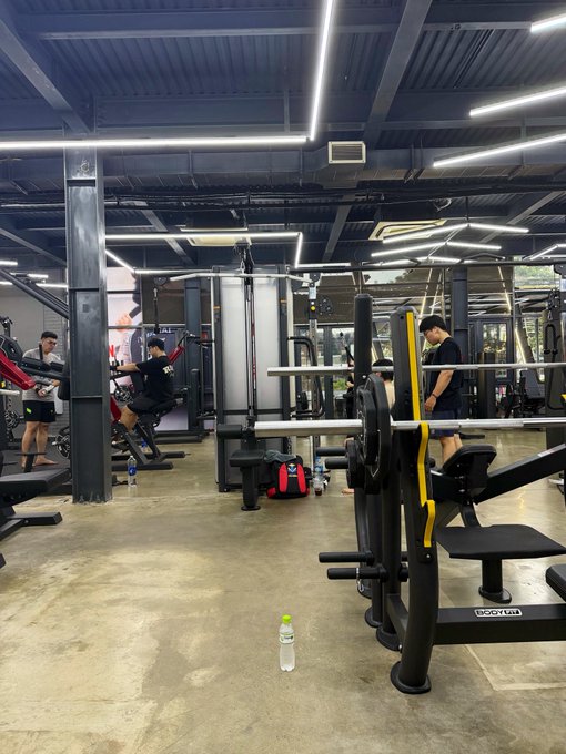 A man lifts weights at an industrial-style gym featuring a BodyMax branded bench press, with several other patrons working out in the background under bright LED ceiling lights. The image's fitness theme pairs with the post's motivational tone about doubling down on crypto bets, both conveying discipline and strength-building through their respective pursuits.