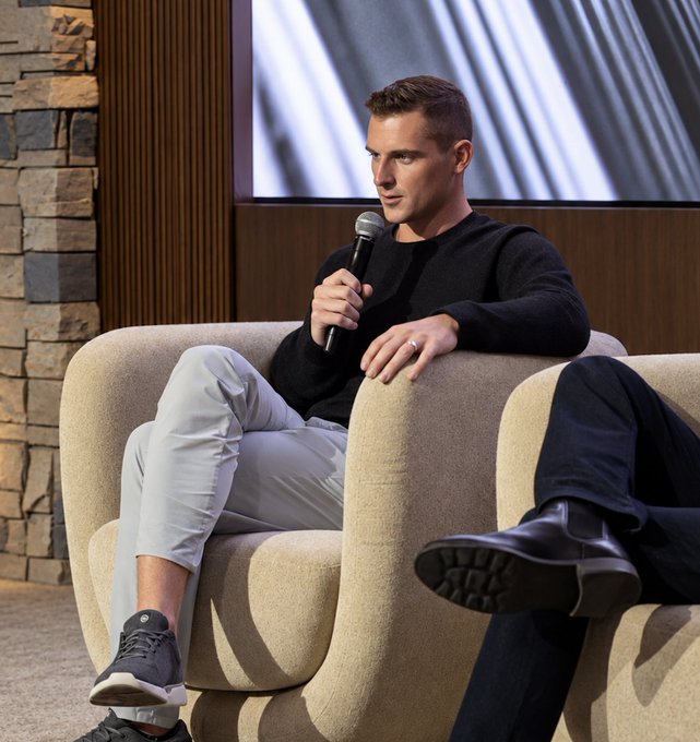 Bo Hines, CEO of Tether U.S., sits casually on a beige armchair holding a microphone during a panel discussion, as he jokes about the surprisingly swift acceptance of blockchain and stablecoins within the U.S. government.