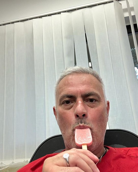 A middle-aged man with gray hair takes a selfie while holding a pink popsicle near his mouth in front of vertical window blinds.