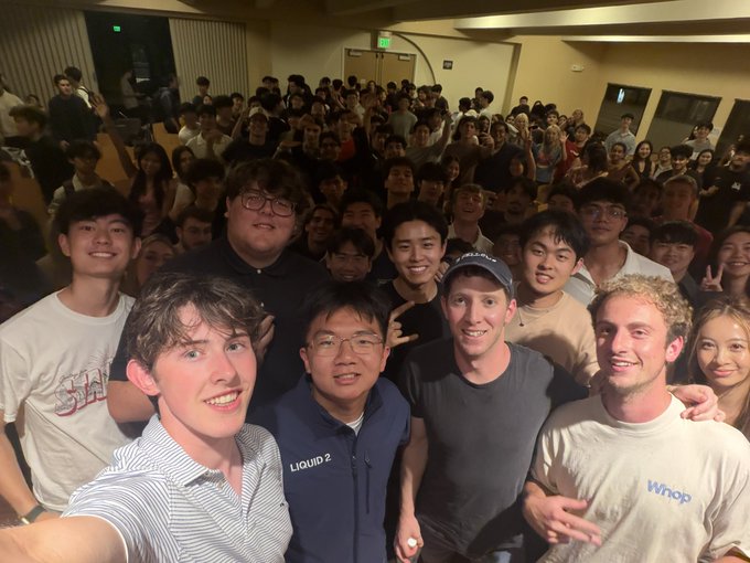 A large group selfie taken at an indoor event, featuring a young man taking the photo alongside several others, including individuals wearing shirts with 'LIQUID 2' and 'whop' branding, with a crowd behind them.