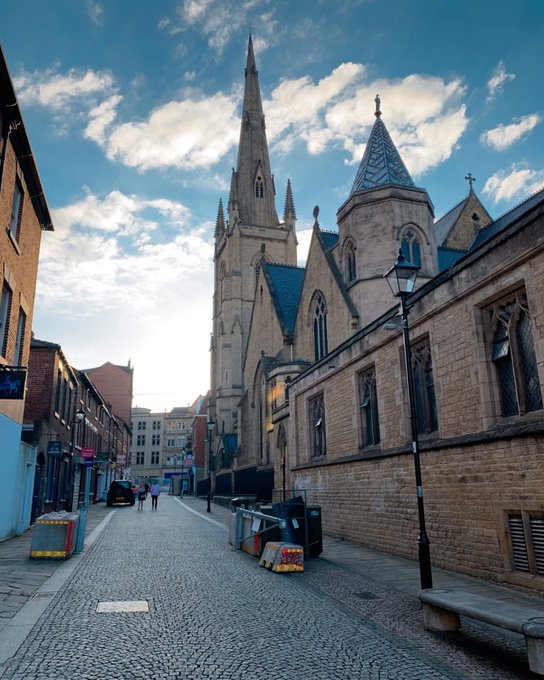 A picturesque cobblestone street lined with historic stone buildings and a Gothic-style church with a tall spire under a partly cloudy blue sky.
