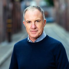 A professional headshot of a middle-aged man with short gray hair, wearing a navy sweater over a striped collared shirt, photographed on a wooden boardwalk.