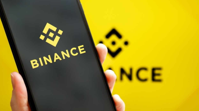 A smartphone displays the yellow Binance logo on its screen, held against a matching yellow background featuring the Binance brand name.