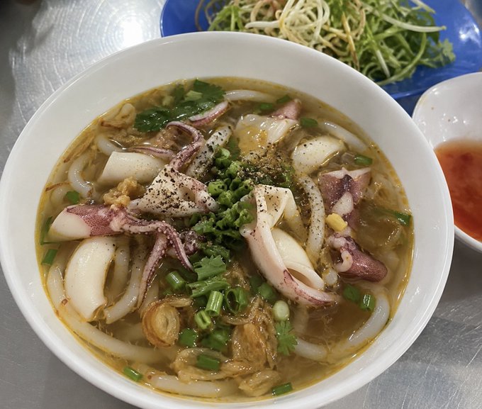 A close-up photo of a bowl of Vietnamese noodle soup, likely bánh canh, topped with squid, green onions, black pepper, and fried shallots, served with a side of bean sprouts and chili sauce.