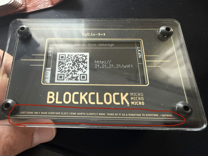 A hand holds a clear acrylic BlockClock MICRO device displaying a Bitcoin seed phrase, QR code, and the URL http://21.21.21.21/uifi, with a circled Satoshi quote at the bottom reading 'LOST COINS ONLY MAKE EVERYONE ELSE'S COINS WORTH SLIGHTLY MORE. THINK OF IT AS A DONATION TO EVERYONE. -SATOSHI'.