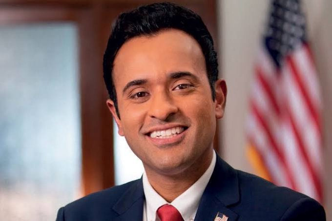 A professional headshot of a smiling man in a dark suit with a red tie, appearing to be a politician, with an American flag visible in the background.