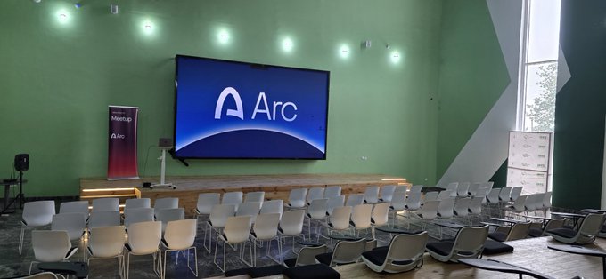 A modern event hall set up for a corporate presentation, featuring rows of empty white chairs facing a large screen displaying the "Arc" logo. To the left, a "MeetUp" banner with the Arc logo reinforces the brand, visually supporting the post's message about Arc building the financial rails for the emerging agentic economy.