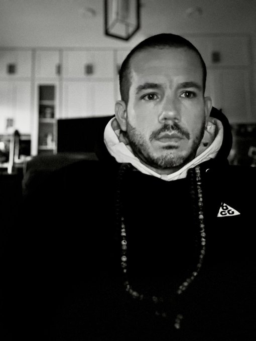 A black and white portrait photo of a bearded man wearing a dark Nike ACG hoodie and beaded necklaces, shot in what appears to be a kitchen setting.