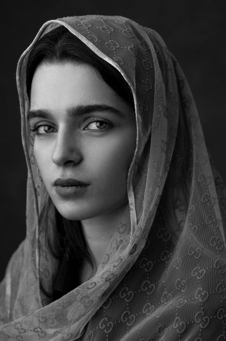 A black-and-white portrait of a young woman wearing a Gucci-patterned veil draped over her head and shoulders.