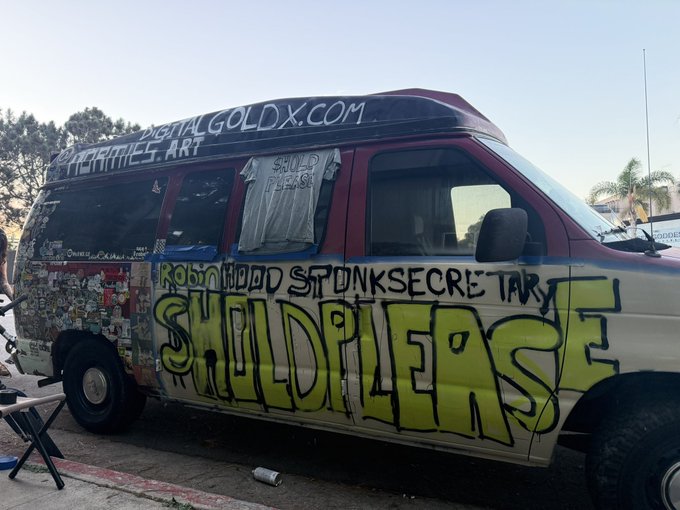 A graffiti-covered van parked on the street featuring prominent yellow 'SHIBPLEASE' artwork, along with tags like 'Robinhood', 'STONKS', and 'SECRETAR' on its side panels.