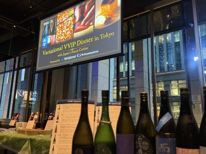 A row of sake bottles is displayed at the "Variational VVIP Dinner in Tokyo" event featuring Japan's finest cuisine, presented by Variational and The Consulting Group, with the celebratory mood of zero trading fees reflected in the elegant reception setting.