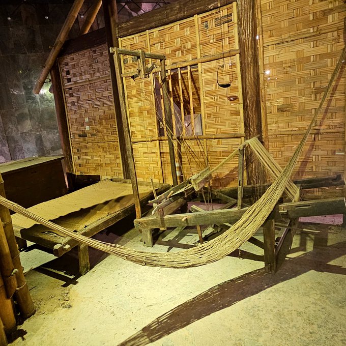 A photograph of a traditional bamboo and wood weaving loom displayed indoors against woven bamboo walls, with loose threads draped over its wooden frame.