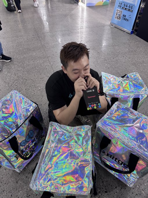 A man kneels on the floor biting open a Bitgear product box, surrounded by several holographic iridescent packaging boxes and bags in what appears to be a store or event space.