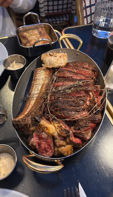 A close-up photo of a large oval pan filled with a sliced tomahawk steak and roasted garlic, served on a dark table alongside small sauce cups.