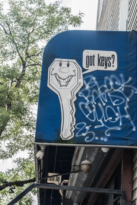 A blue sign on a building features a cartoon key with a smiling face and a speech bubble reading 'got keys?', partially covered in white graffiti.