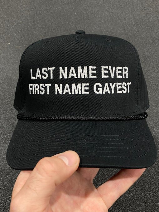 A hand holding a black baseball cap with white embroidered text reading: 'LAST NAME EVER, FIRST NAME GAYEST'.