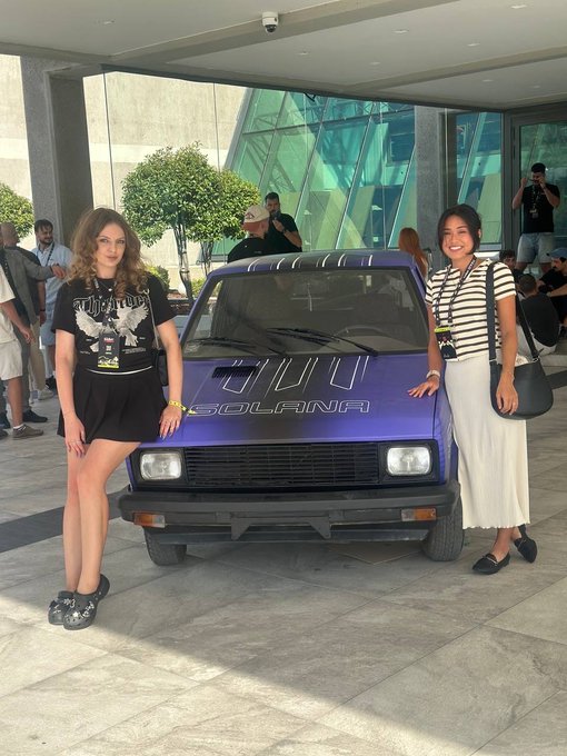 Two women posing on either side of a purple car with 'SOLANA' and a logo written on the hood, parked outside a modern building with glass and geometric patterns; one woman wears a black t-shirt and skirt, the other a striped shirt and white skirt.