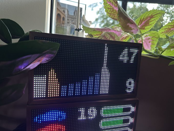 An LED dot matrix display showing colorful graphics including a rocket ship and the numbers '11', '7', '2', and '5', situated near a window with plants.