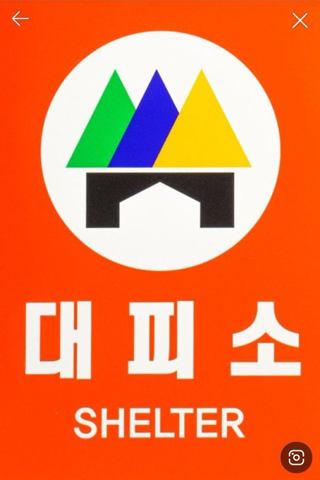 The photo shows an official South Korean civil defense shelter sign with a red background, displaying the Korean text "대피소" (Shelter) and English "SHELTER," featuring a logo with three colored triangular peaks in green, purple (blue), and yellow. The caption draws a playful parallel between these shelter alert colors and the cryptic "BearableGuy" riddles circulating in crypto circles, which assign BTC to green, XRP to purple, and USD to yellow — implying the colors themselves might be signaling an XRP pump.