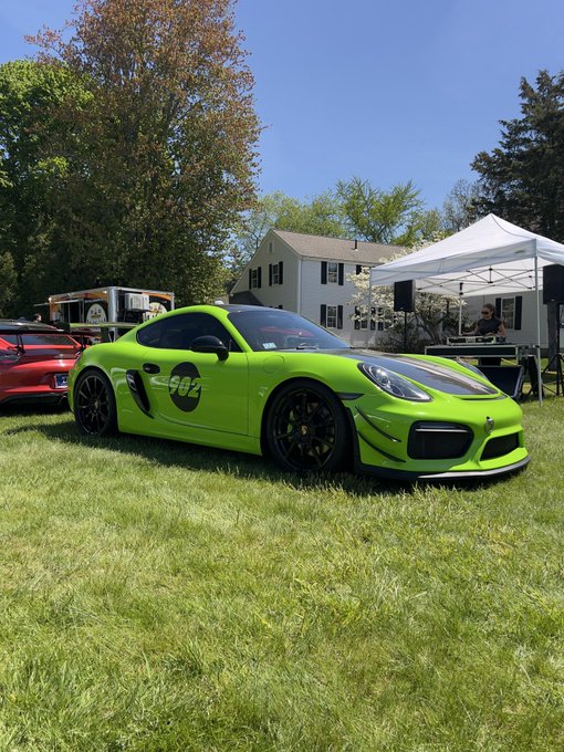 A bright green Porsche Cayman with "992" racing decals and a black hood is parked on grass at what appears to be a car show, with a white tent and other vehicles visible in the background.