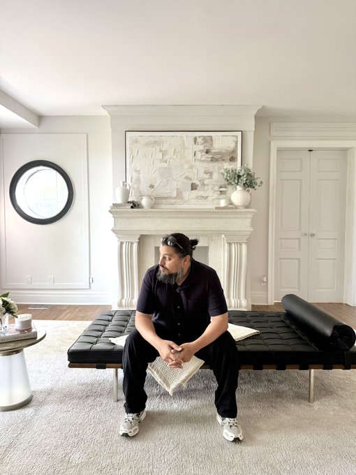 A bearded man with a topknot, wearing a dark button-down shirt, sits on a black leather Barcelona bench in a minimalist white living room with a fireplace and round mirror. The caption suggests he's preparing to sleep and will select a subscriber for a vouch giveaway upon waking, though no text or names are visible in the image itself.