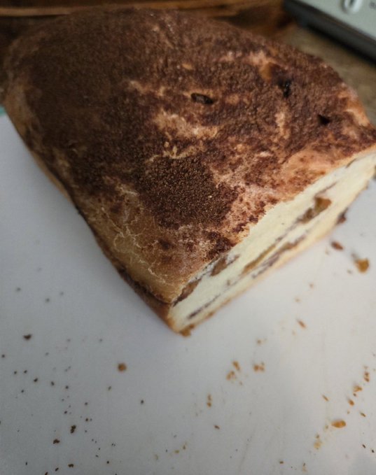 A rectangular slice of tiramisu with a cocoa-dusted top sits on a white cutting board with crumbs scattered around.