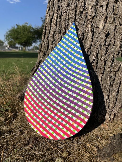 A large teardrop-shaped object featuring a colorful checkered pattern in pink, blue, green, and purple, leaning against the trunk of a tree in a grassy park.