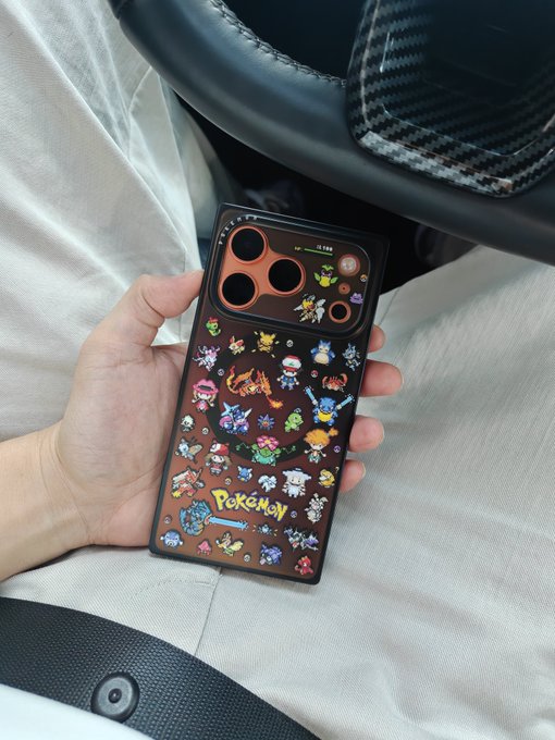 A hand holds a smartphone encased in a brown cover featuring numerous Pokémon stickers and the 'Pokémon' logo, resting over beige pants near a car seat.