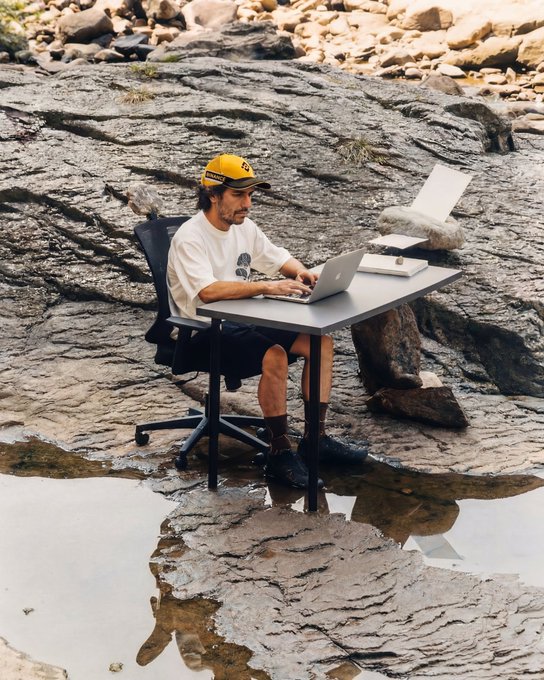 A man wearing a yellow cap and white t-shirt works at his laptop while seated in a black office chair with a makeshift desk, set up on rocky terrain beside a reflective pool of water, embodying the rugged yet remote "latest setup" of outdoor productivity.