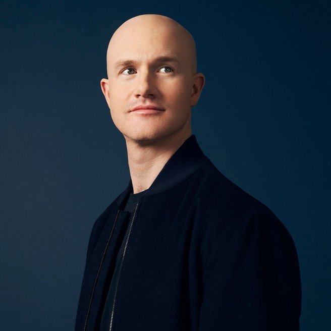 A bald man with light-colored eyes wears a dark navy bomber jacket over a black shirt, posing against a solid dark blue background.