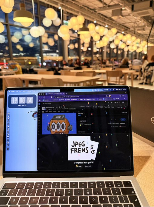 A laptop on a table in a cafe displaying a trading interface with 'JPEG FRENS' text and an NFT image of a cartoon character, with the clock showing 19:00:50.