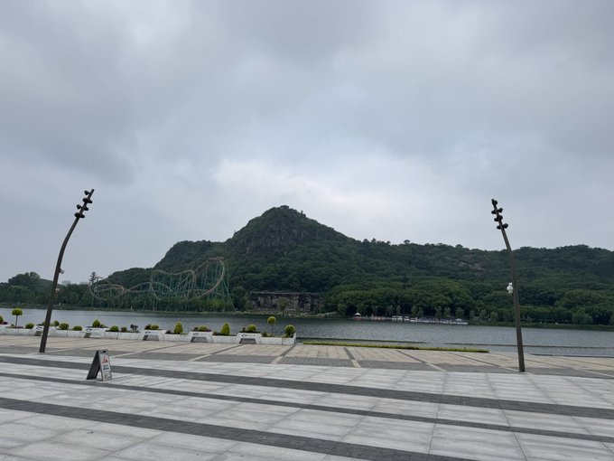 The image shows a large, gray stone structure with a mountain range in the background. The sky above appears cloudy, creating an overcast atmosphere. In front of this scene, there are two street lamps standing side by side, one on each end of the street. A person is also visible near these street lamps, possibly walking or waiting for someone else to arrive.